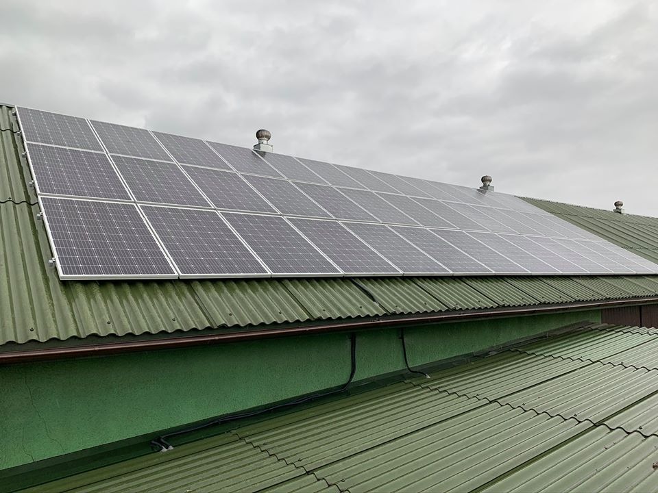 Instalacja paneli słonecznych na zielonym, falistym dachu z blachy, widoczne przewody elektryczne i wentylatory dachowe przy pochmurnym niebie.