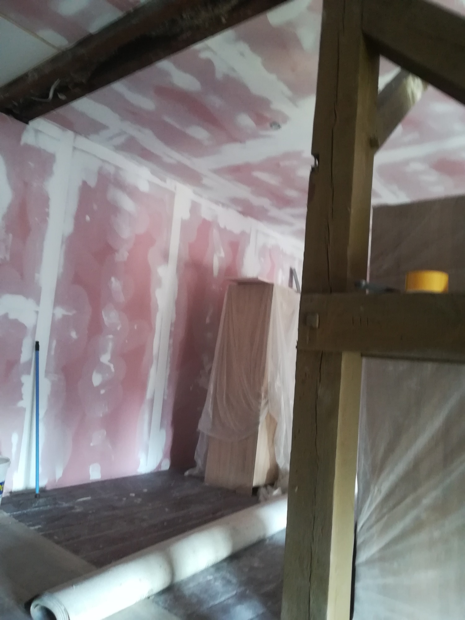 Pokój w trakcie remontu: ściany pokryte różowym podkładem i częściowo szpachlowane, widoczne stare drewniane belki stropowe i rolka materiału podłogowego.