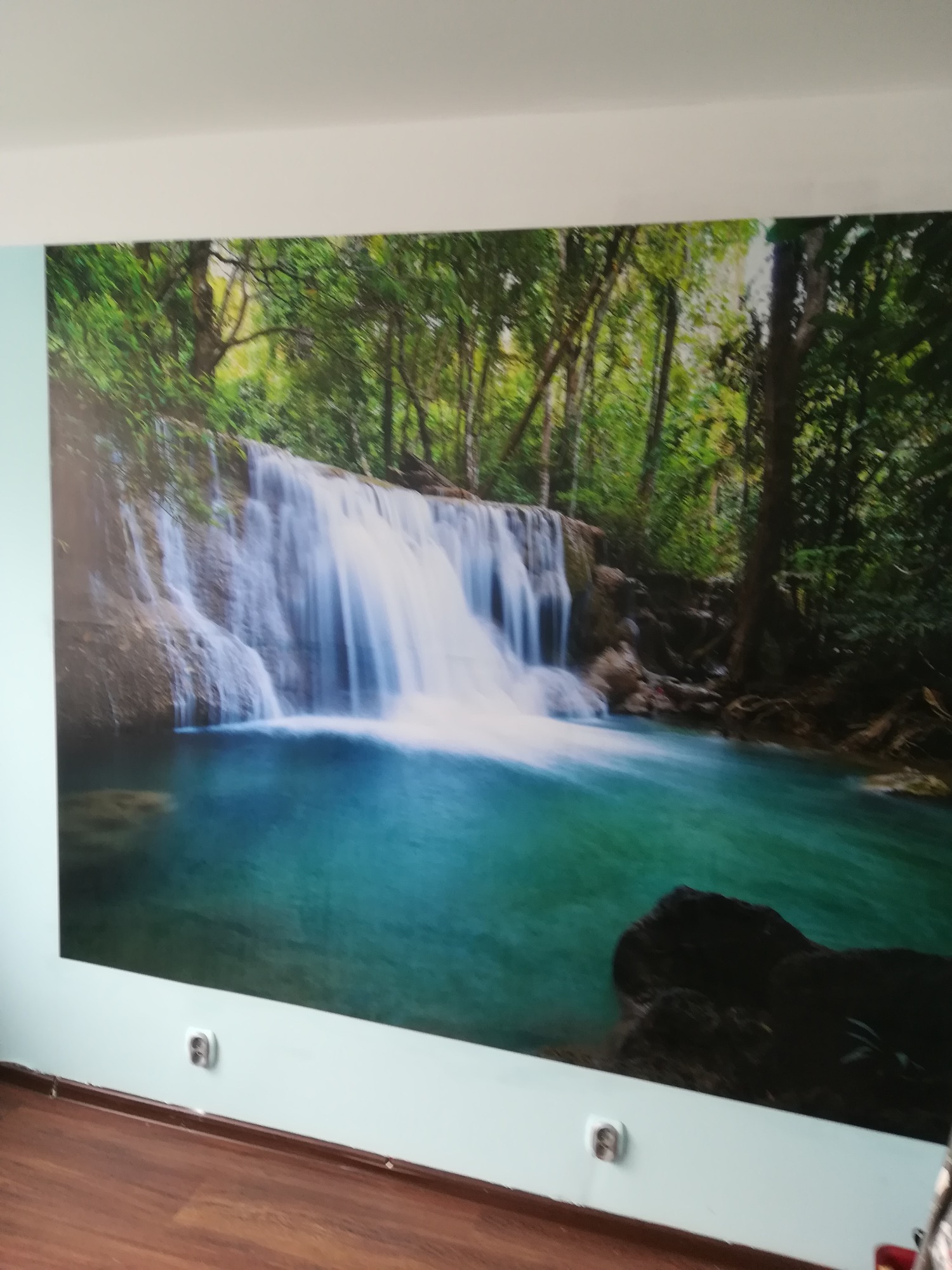 Fototapeta z motywem wodospadu w dżungli, zamontowana na ścianie w pokoju z brązową podłogą, widoczne gniazdka elektryczne.