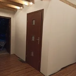 Wnętrze z drewnianą podłogą imitującą panele, białe ściany, drewniane belki stropowe z żarówkami i brązowe drzwi z małymi szybkami.