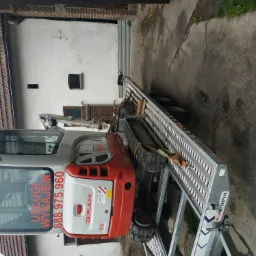 Mini koparka Takeuchi TB216 na metalowej przyczepie transportowej z widocznym numerem telefonu kontaktowego i napisem 'Usługi Wynajem' na szybie, zaparkowana na betonowym podwórku przed budynkiem.