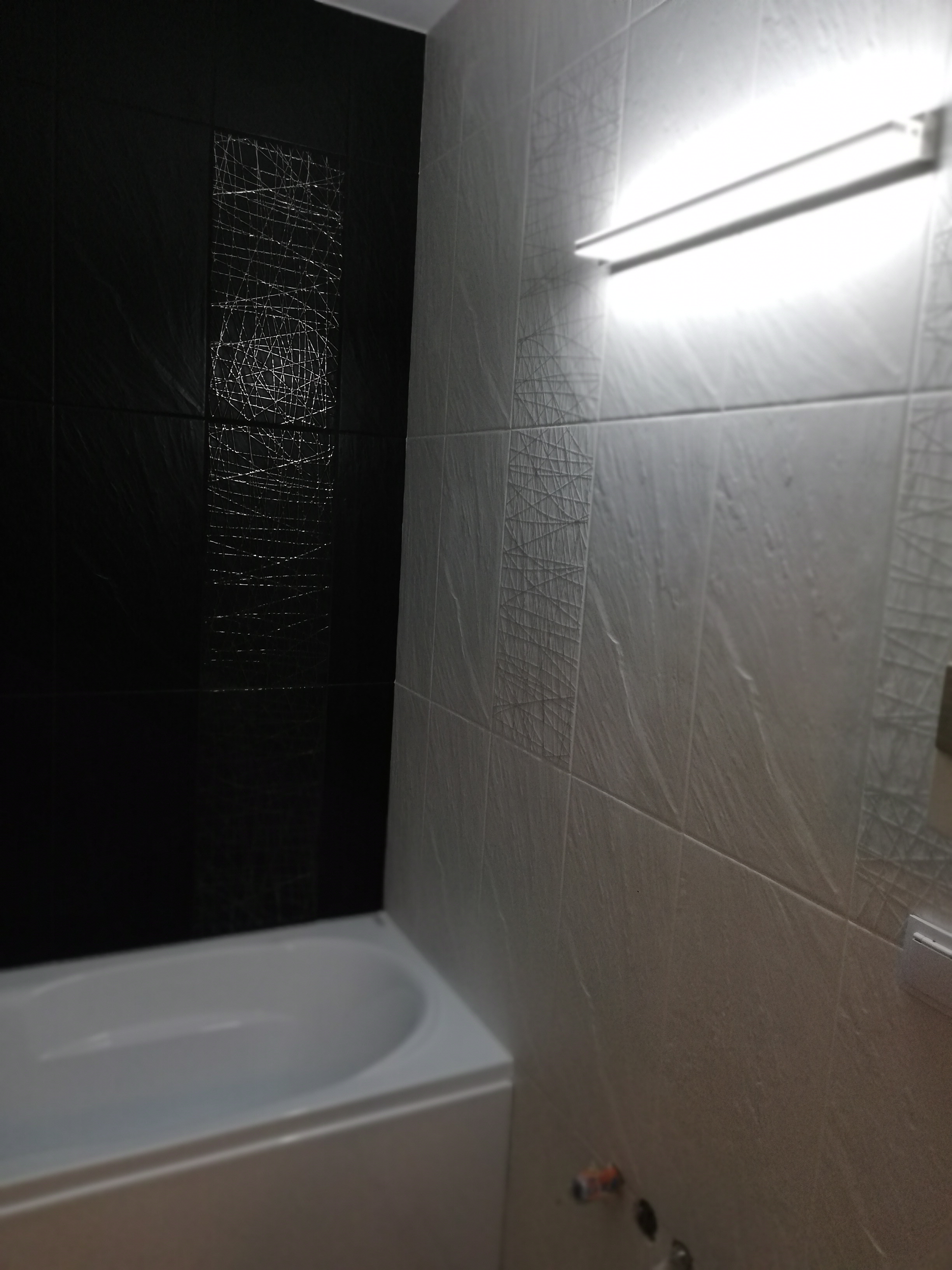 Nowoczesna łazienka z czarno-białymi płytkami o wzorze przypominającym poplątane linie, oświetlona podłużną lampą nad wanną.