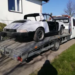 Uszkodzony, częściowo odmalowany Porsche Targa na platformie białej lawety, widoczny numer telefonu na kabinie pojazdu holowniczego, dom w tle.