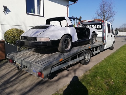 Uszkodzony, częściowo odmalowany Porsche Targa na platformie białej lawety, widoczny numer telefonu na kabinie pojazdu holowniczego, dom w tle.