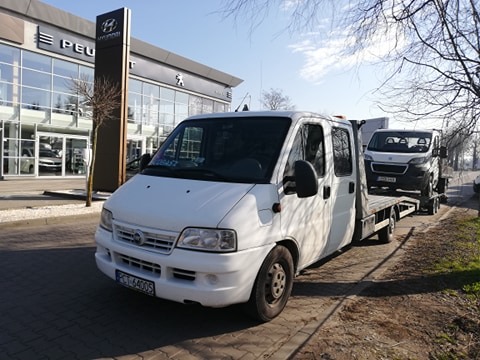 Biała laweta z załadowanym białym busem na tle salonu samochodowego Hyundai i Peugeot.