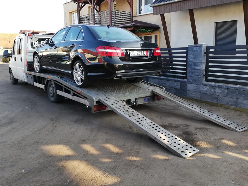 Czarny Mercedes E-klasa z numerem rejestracyjnym OE 257 E na platformie białej lawety z rozłożonymi rampami, zaparkowanej na żwirowym podłożu przed domem.