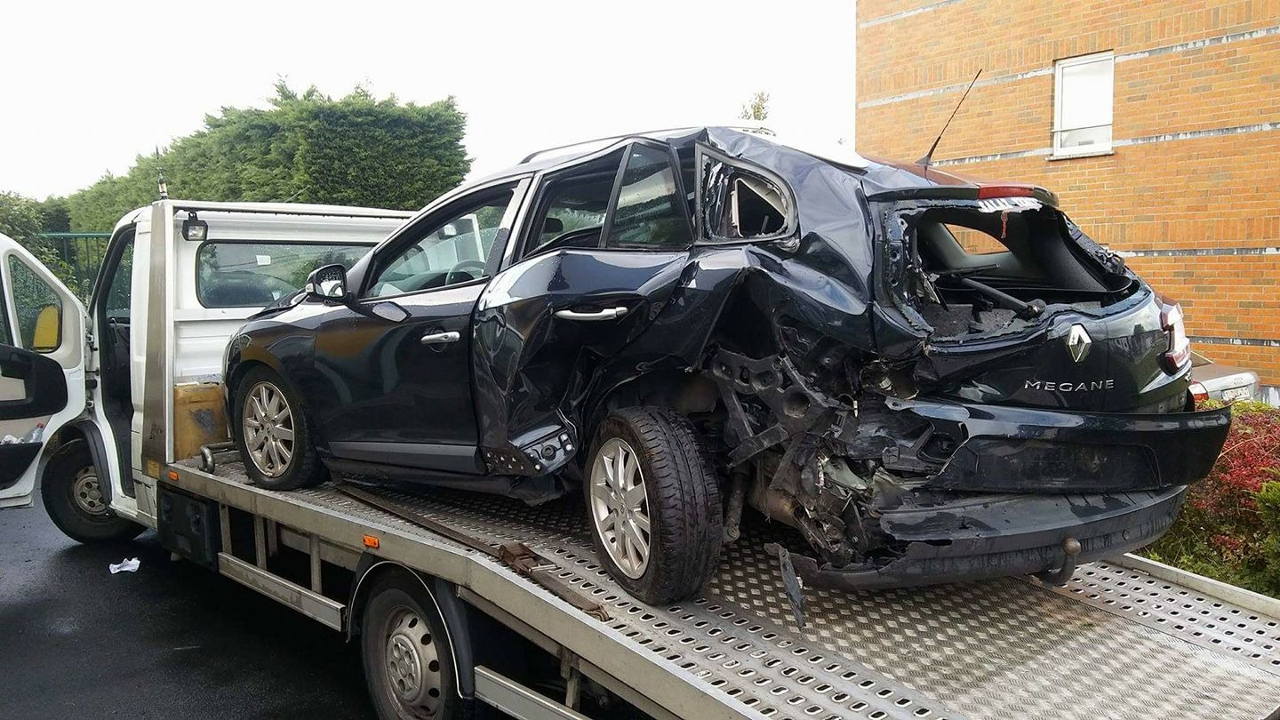 Czarny Renault Megane po wypadku, z poważnymi uszkodzeniami karoserii, umieszczony na platformie białej lawety, na tle ceglanego budynku i zieleni.