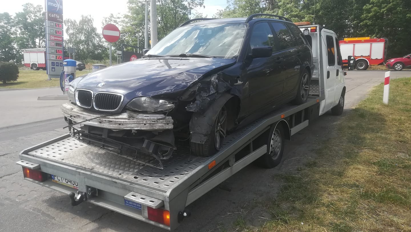 Uszkodzony, granatowy samochód osobowy BMW kombi załadowany na platformę lawety. W tle widoczna stacja paliw Moya i wóz strażacki.