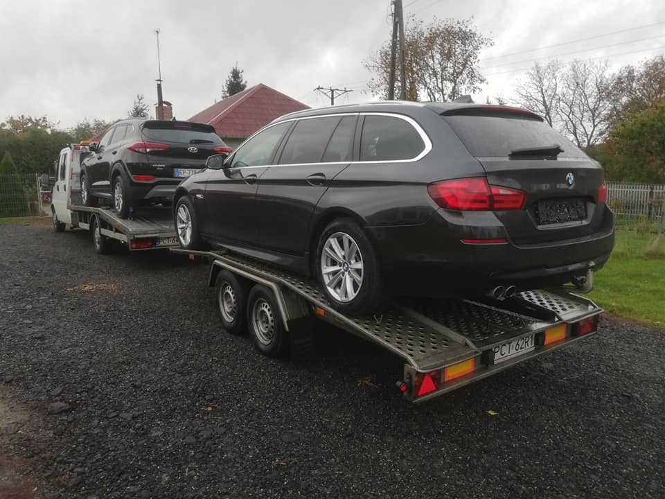 Dwa samochody, czarny Hyundai Tucson i ciemnoszary BMW kombi, przewożone na dwóch przyczepach przez biały samochód dostawczy w 64-720. Widok z tyłu na BMW, z widocznymi tablicami rejestracyjnymi...