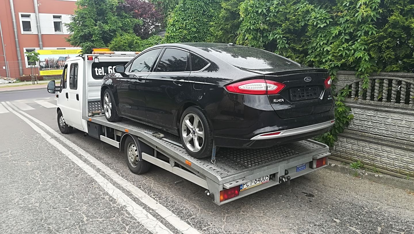 Czarny Ford Riata zabezpieczony na platformie autolawety, widok z tyłu, na tle zieleni i budynku.