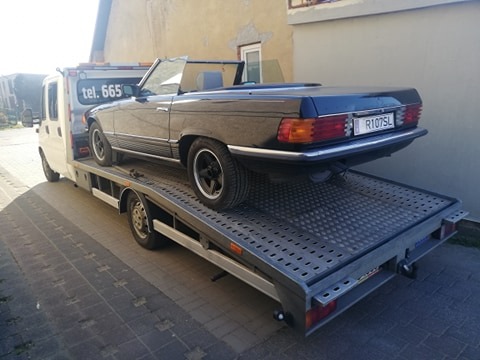 Czarny kabriolet Mercedes-Benz na platformie lawety, widok z boku, budynek w tle.