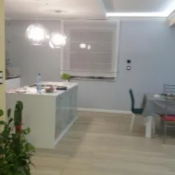 Nowoczesna kuchnia z wyspą, drewnopodobną podłogą, oświetleniem LED w suficie i wiszącymi lampami kulami, z widocznym stołem jadalnym w tle.