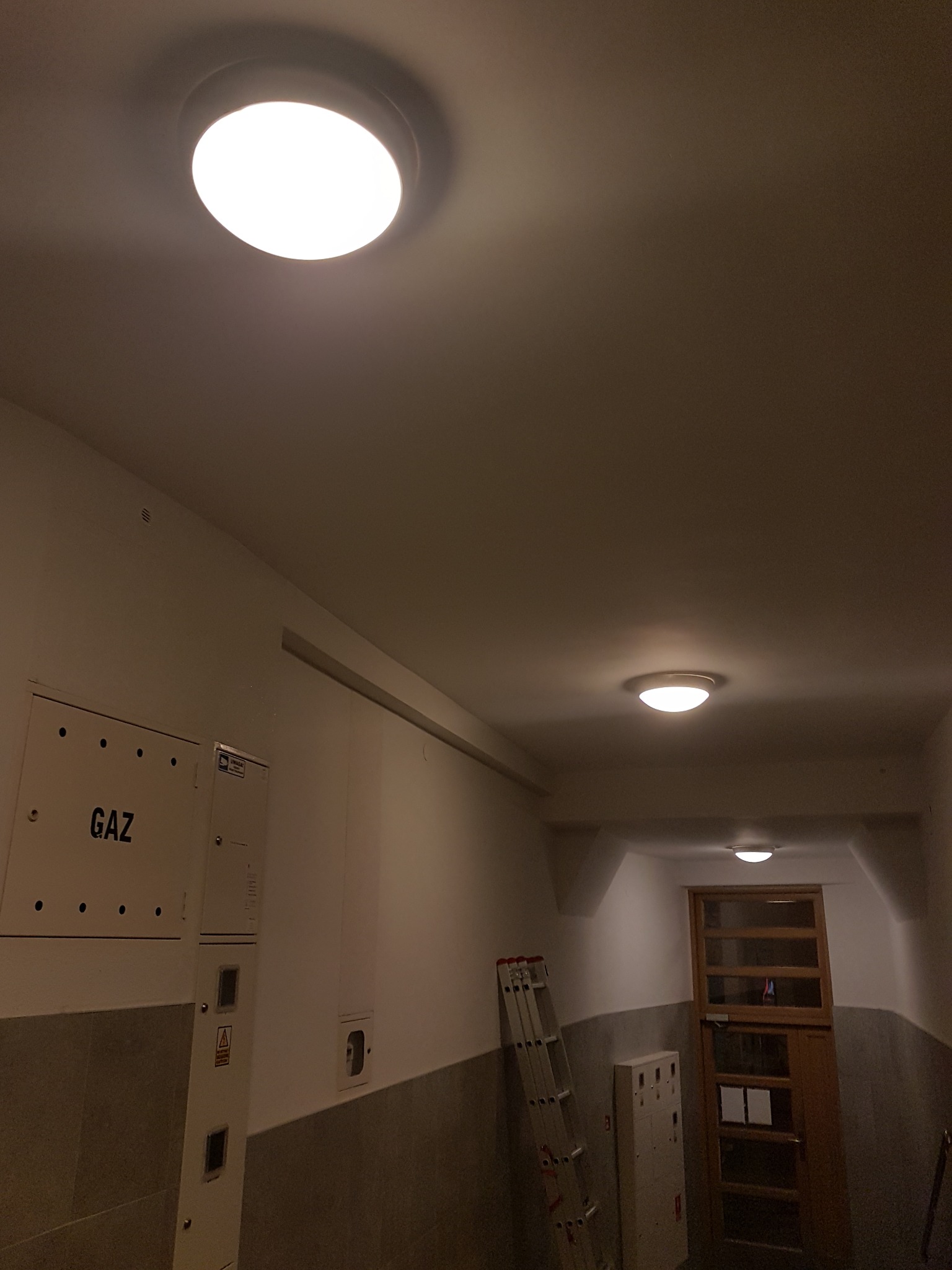 Korytarz w bloku mieszkalnym z trzema okrągłymi lampami sufitowymi, skrzynką gazową i metalową drabinką opartą o ścianę.