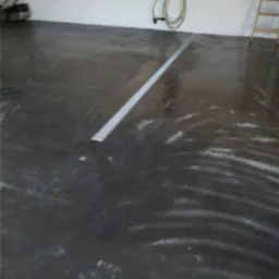 Błyszcząca, ciemnoszara posadzka przemysłowa z odpływem liniowym w garażu lub hali, widoczna kosiarka i drabina w tle.