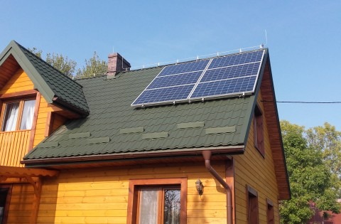 Żółty dom jednorodzinny z ciemnozielonym dachem, na którym zamontowano sześć paneli słonecznych. Widoczny komin z cegły i antena na dachu. Błękitne niebo w tle.