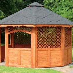 Drewniana altana ogrodowa z sześciokątnym dachem krytym gontem bitumicznym, z ozdobnymi kratami i panelami, otoczona zielonym trawnikiem i drzewami.