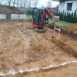 Mini koparka na placu budowy, przygotowany wykop pod fundamenty z widocznymi białymi oznaczeniami, w tle hałda ziemi i budynek mieszkalny.