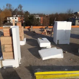 Wznoszenie muru z bloczków ceramicznych Porotherm i białych elementów izolacyjnych na dachu budynku, widoczne narzędzia i materiały budowlane, w tle zabudowa mieszkalna i drzewa.