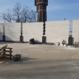 Wznoszenie muru z białych bloczków betonowych, widoczne zbrojenie w pionowych szczelinach, w tle wieża ciśnień i błękitne niebo.