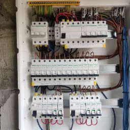 Rozdzielnica elektryczna z zamontowanymi bezpiecznikami i uporządkowanymi przewodami w białej obudowie, zamocowana na surowej ścianie z widocznymi śladami tynku i notatkami.