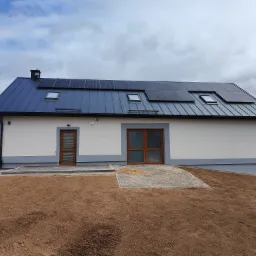 Nowoczesny dom jednorodzinny z panelami słonecznymi na dachu z blachy na rąbek stojący i oknami dachowymi, świeżo wyrównany teren przed budynkiem.