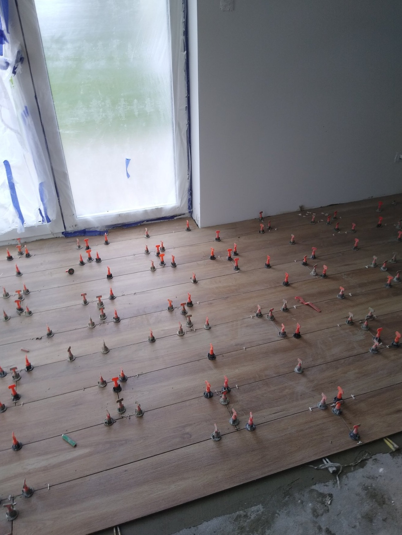 Układanie paneli podłogowych imitujących drewno, z użyciem pomarańczowych klinów dystansowych, widoczne zabezpieczone okno folią malarską i fragment ściany w surowym stanie.