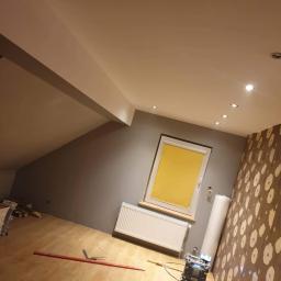 Gol-Bud - Pokój na poddaszu w trakcie remontu: szare ściany, wzorzysta tapeta, okno z żółtą roletą, grzejnik, narzędzia do układania paneli i rolka tapety przy ścianie.