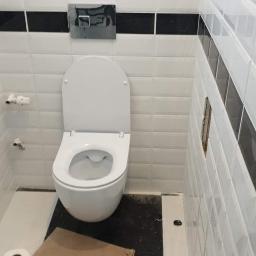 Gol-Bud - Biała toaleta z otwartą klapą w wykończonej łazience z białymi płytkami z czarnymi paskami, pod toaletą leży kawałek kartonu i rolka papieru toaletowego.