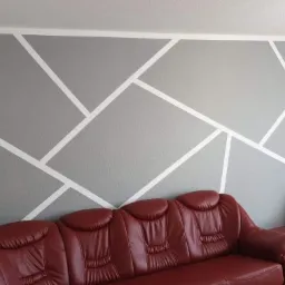 Szara ściana z geometrycznym wzorem z białych pasów, uzupełniona o czerwoną skórzaną sofę i głośniki po bokach, tworząca nowoczesną aranżację wnętrza.