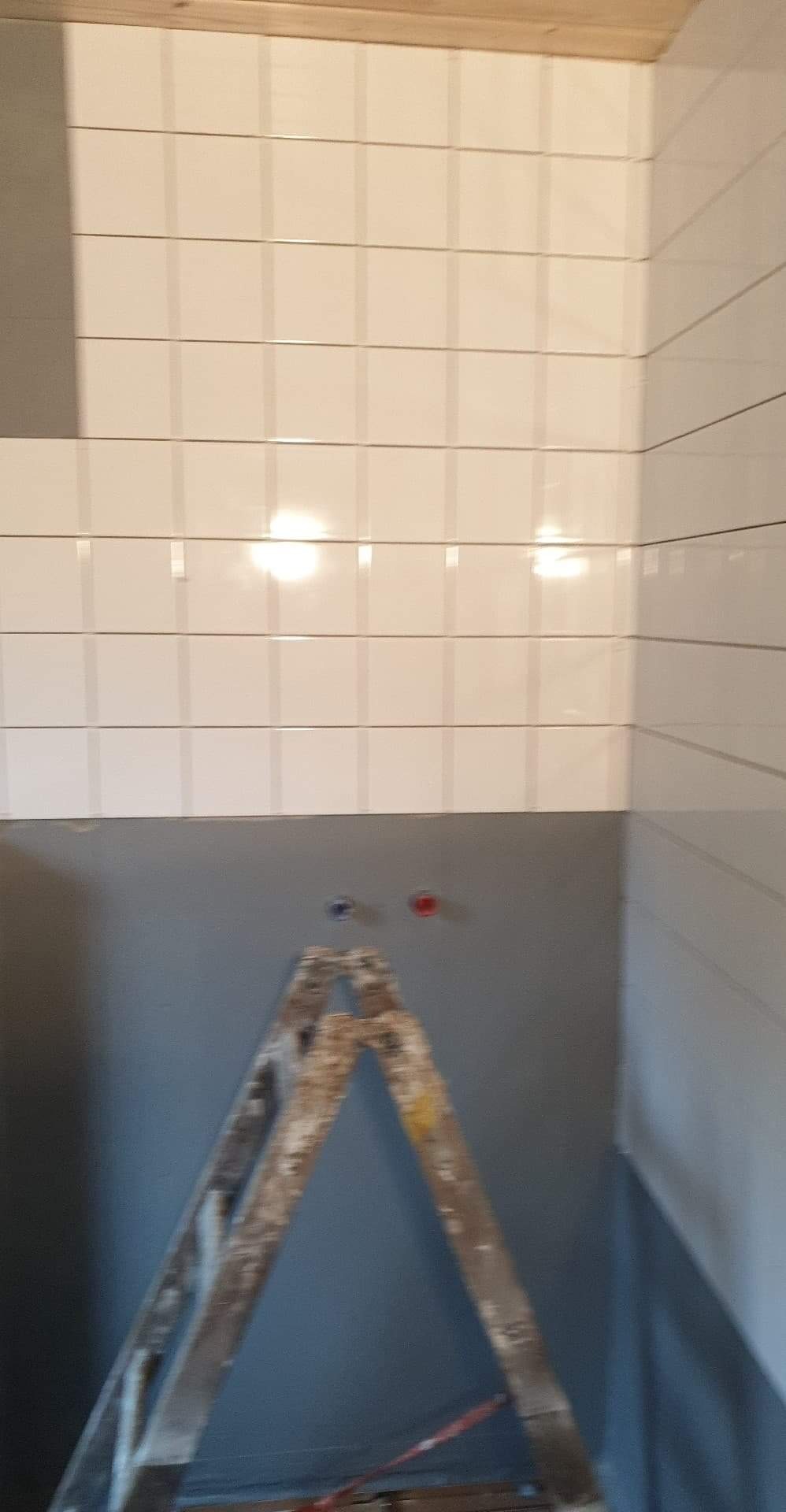 Ściana w łazience w trakcie wykańczania, z białymi, kwadratowymi płytkami ceramicznymi ułożonymi do pewnej wysokości, poniżej szara ściana przygotowana pod malowanie, widoczna drabina malarska...