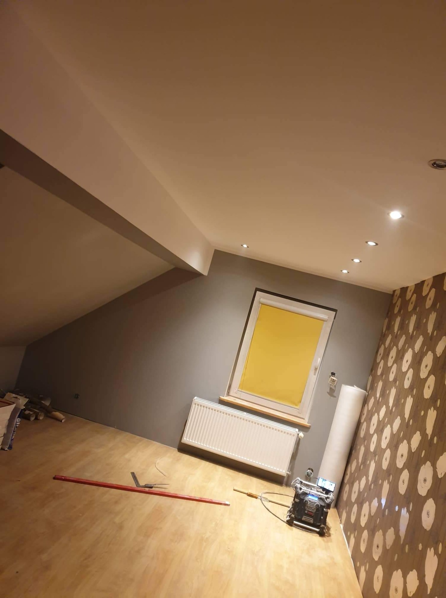 Pokój na poddaszu w trakcie remontu: szare ściany, wzorzysta tapeta, okno z żółtą roletą, grzejnik, narzędzia do układania paneli i rolka tapety przy ścianie.
