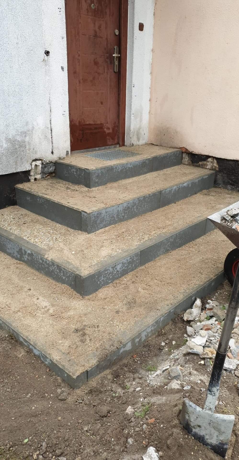 Schody wejściowe z szarych prefabrykatów, zasypane piaskiem, z widoczną łopatą i gruzem, prowadzące do brązowych drzwi.