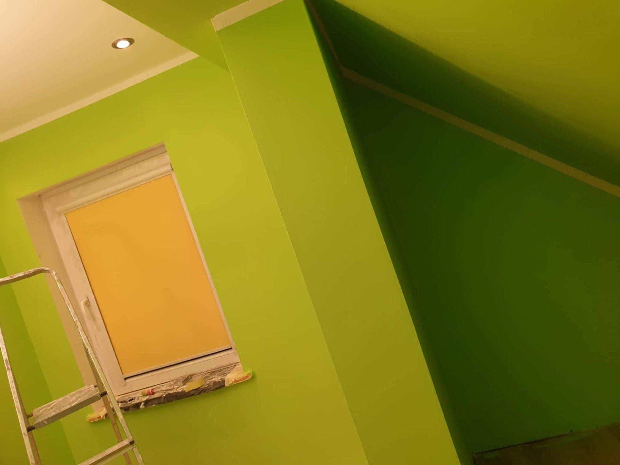 Świeżo pomalowane na jasnozielono poddasze ze skosami, okno z zasłoniętą roletą zabezpieczone taśmą malarską, fragment drabiny malarskiej.