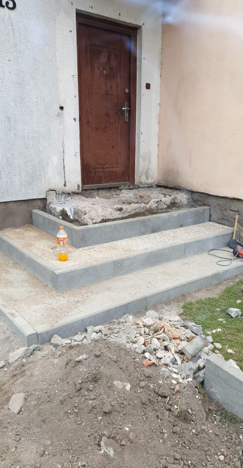 Nowe betonowe schody wejściowe z szarą kostką ułożoną na stopniach, w trakcie budowy, z butelką napoju na jednym ze stopni, obok stare, zniszczone schody.
