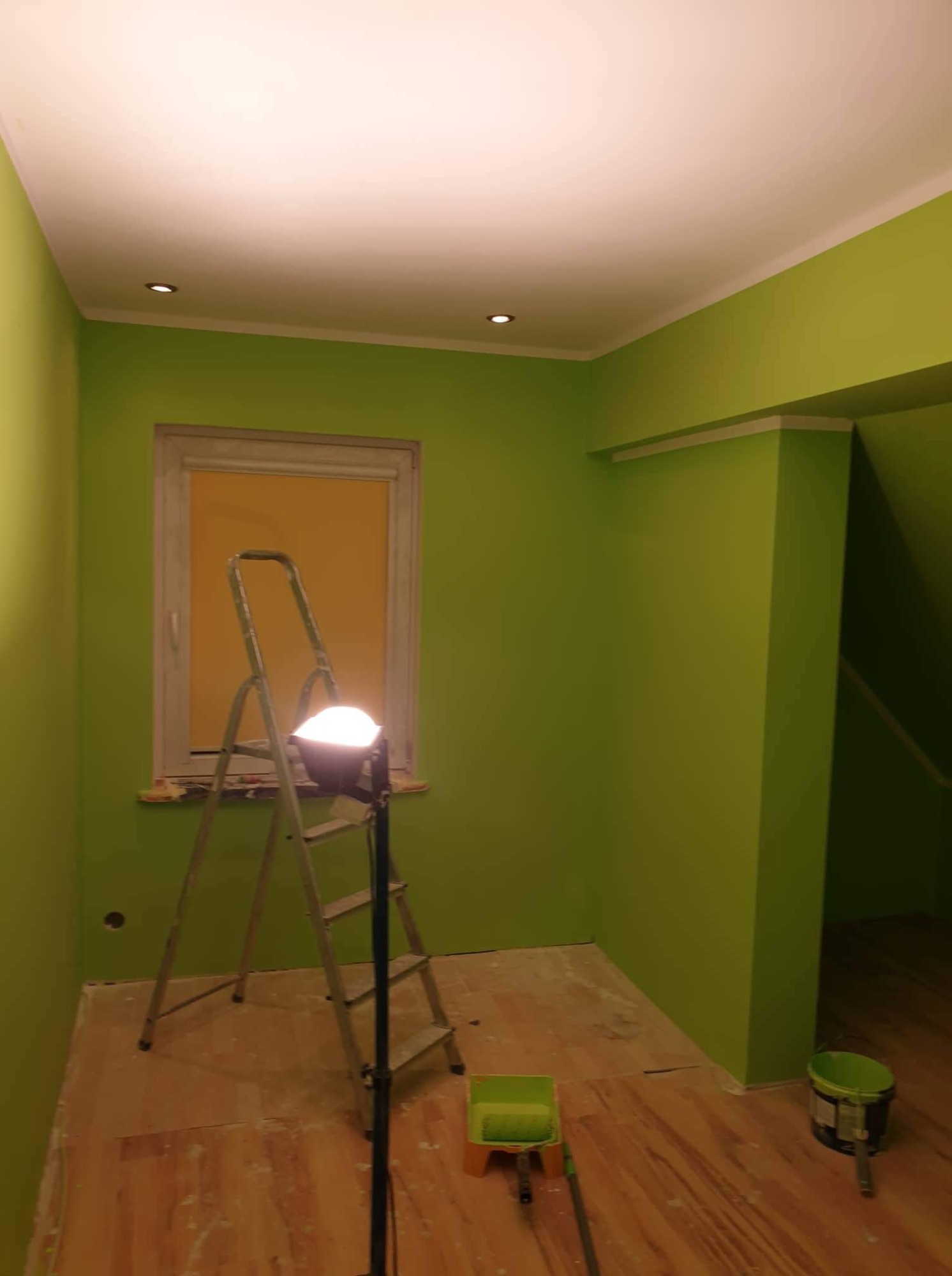 Świeżo pomalowane na zielono wnętrze pokoju z drabiną opartą o okno, lampą budowlaną, widoczne ślady farby na podłodze i wiaderko z farbą.