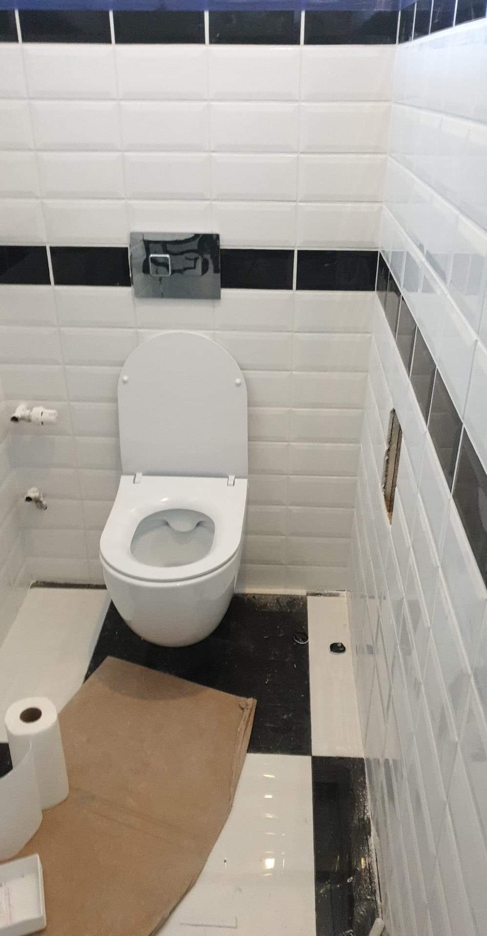 Biała toaleta z otwartą klapą w wykończonej łazience z białymi płytkami z czarnymi paskami, pod toaletą leży kawałek kartonu i rolka papieru toaletowego.
