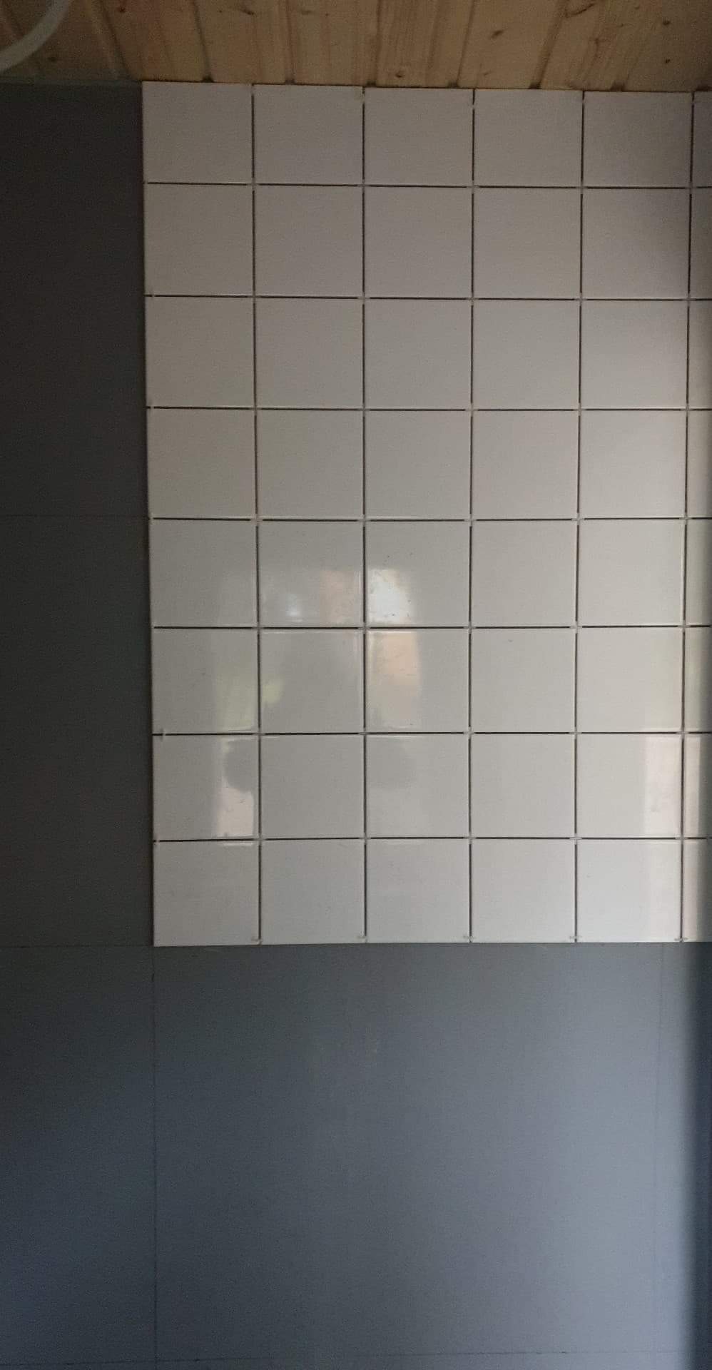 Ściana wyłożona białymi, kwadratowymi płytkami ceramicznymi z ciemną fugą, sąsiadująca z szarą ścianą pomalowaną farbą, widoczne drewniane elementy sufitu.