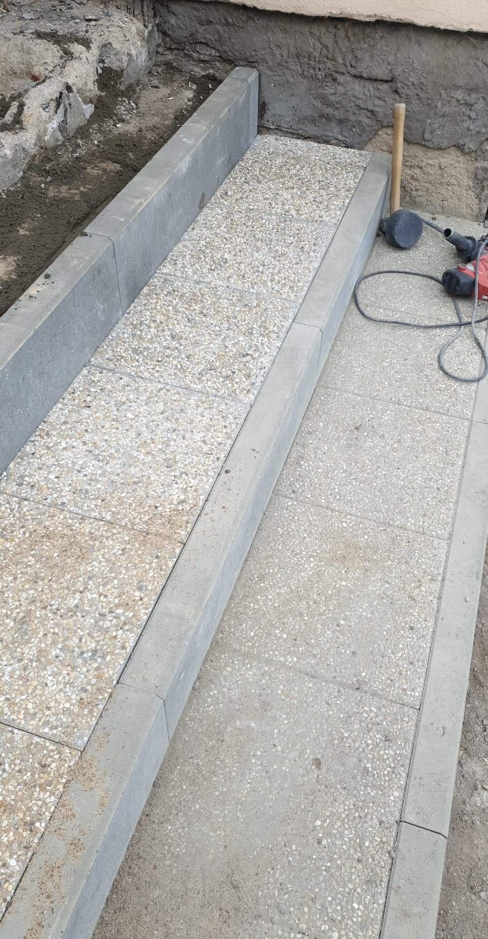 Wykończone schody zewnętrzne z betonowych płyt z drobnym grysem, obrzeża z szarego betonu, widoczne narzędzia budowlane obok.