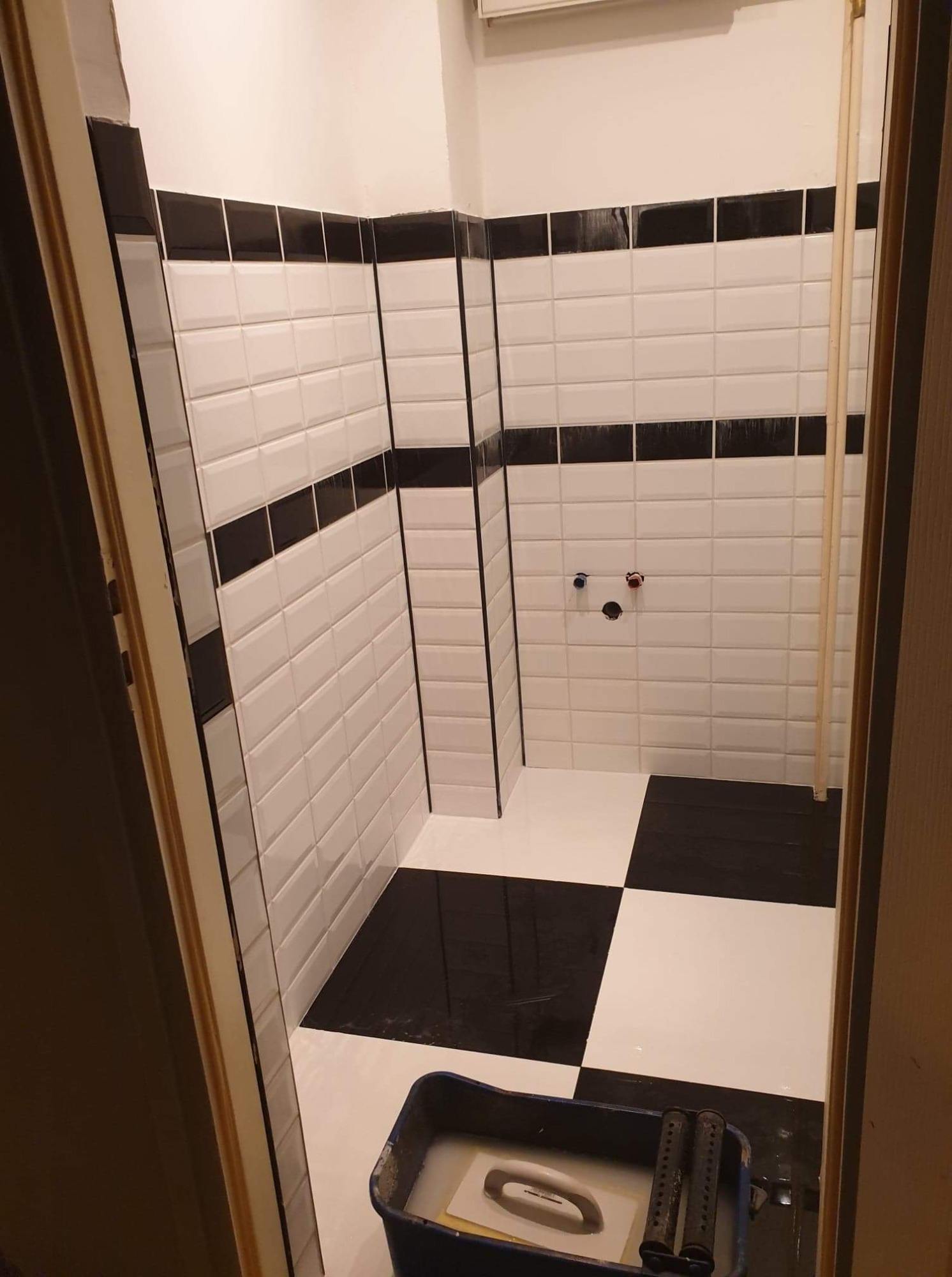 Czarno-biała łazienka w trakcie wykańczania: ułożone płytki w stylu retro, widoczny pojemnik z narzędziami glazurniczymi, fragment rury i przygotowane przyłącza wodne.