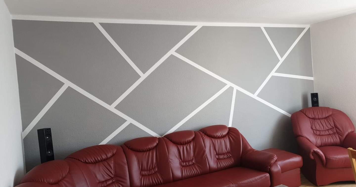Szara ściana z geometrycznym wzorem z białych pasów, uzupełniona o czerwoną skórzaną sofę i głośniki po bokach, tworząca nowoczesną aranżację wnętrza.