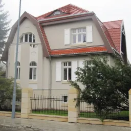 Elewacja dwupiętrowego domu z czerwonym dachem i białymi okiennicami, otoczona niskim kamiennym ogrodzeniem z metalowymi przęsłami i zielenią.