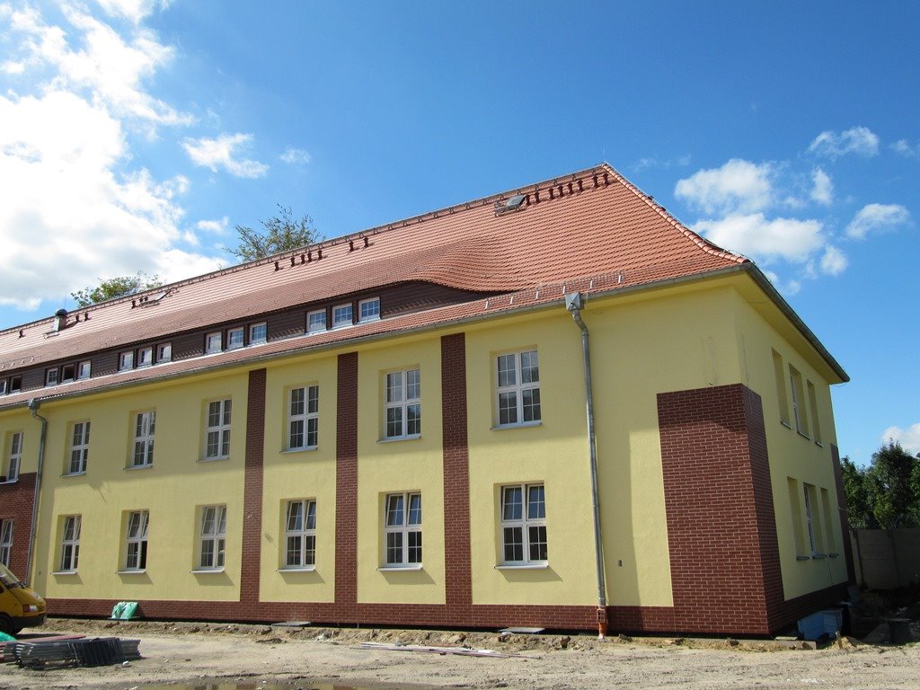 Żółty budynek z czerwonymi ceglanymi detalami i czerwoną dachówką, widoczny z zewnątrz w słoneczny dzień.