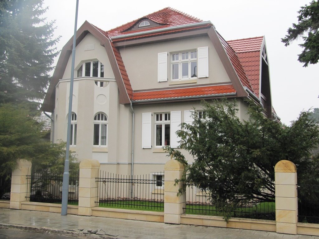 Elewacja dwupiętrowego domu z czerwonym dachem i białymi okiennicami, otoczona niskim kamiennym ogrodzeniem z metalowymi przęsłami i zielenią.