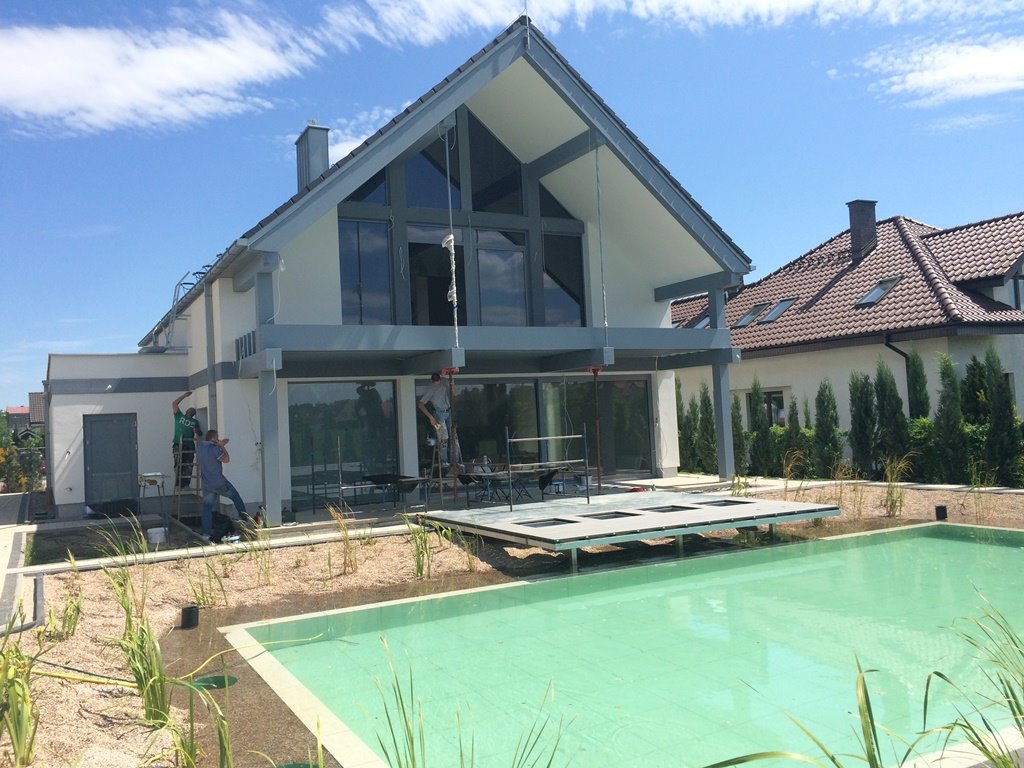 Nowoczesny dom w trakcie wykończenia z basenem, pracownicy na drabinach malują elewację na tle błękitnego nieba.