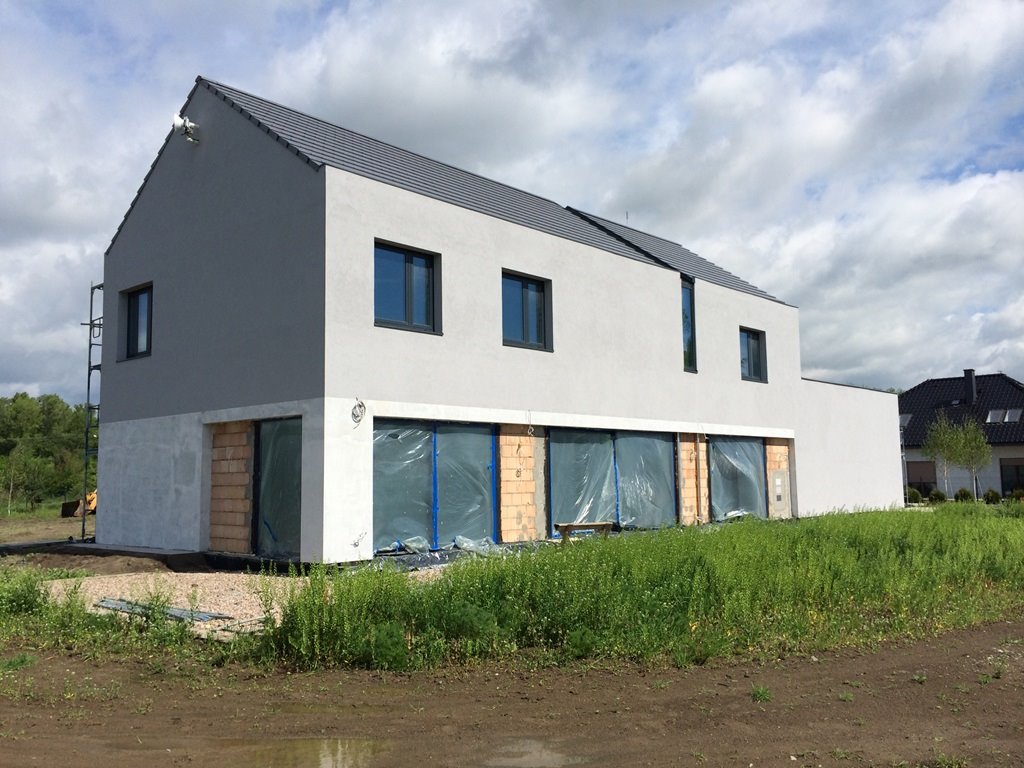 Nowoczesny dom jednorodzinny w trakcie budowy, widoczne okna zabezpieczone folią, surowy stan dolnej części elewacji i niezagospodarowany teren wokół budynku.