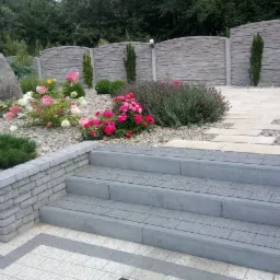 Ogród z kamienną ścieżką, schodami z kostki, rabatami kwiatowymi z różnorodną roślinnością i ozdobnym kamieniem na tle betonowego ogrodzenia.