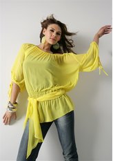 Modelka prezentuje żółtą, zwiewną bluzkę z szerokimi rękawami i wiązaniem w pasie, w połączeniu z dżinsami i srebrną biżuterią na tle białej ściany.