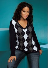 Kobieta o ciemnych, kręconych włosach pozuje w swetrze w romby (argyle) w odcieniach szarości i bieli, z dekoltem w serek, ubrana w ciemne jeansy na tle turkusowej ściany.