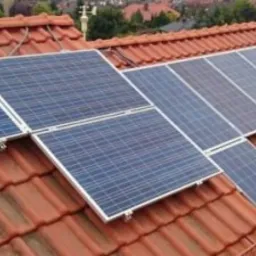 Panele słoneczne zamontowane na dachu z czerwonej dachówki, widok z lekkiego kąta, w tle zabudowa mieszkalna.