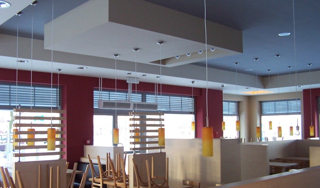 Wnętrze biura z czerwonymi ścianami, żaluzjami w oknach, wiszącymi lampami w odcieniach żółci i pomarańczy oraz podwieszanym sufitem z oświetleniem punktowym.